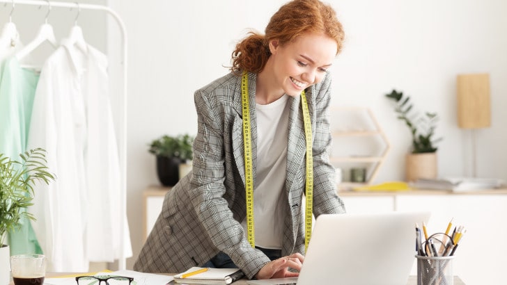 Dressmaker smiles while looking at sewing blogs on her laptop.