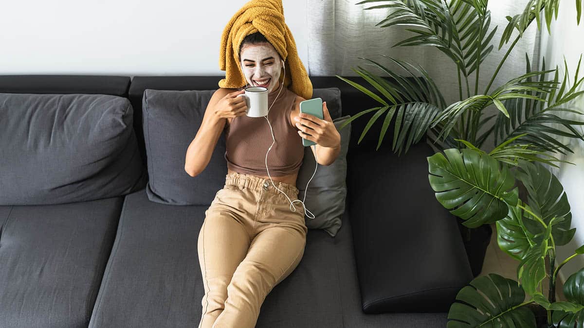 Woman wearing DIY beauty mask at home sips coffee and smiles at her smartphone.