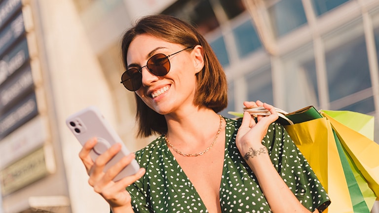 24 Easy Ways to Save Money on Clothes 11 Smiling, stylish shopper stands outside holding smartphone.